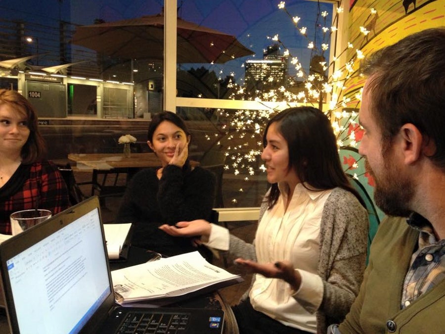 4 people converse while sitting around a table. A laptop and papers are on the table.