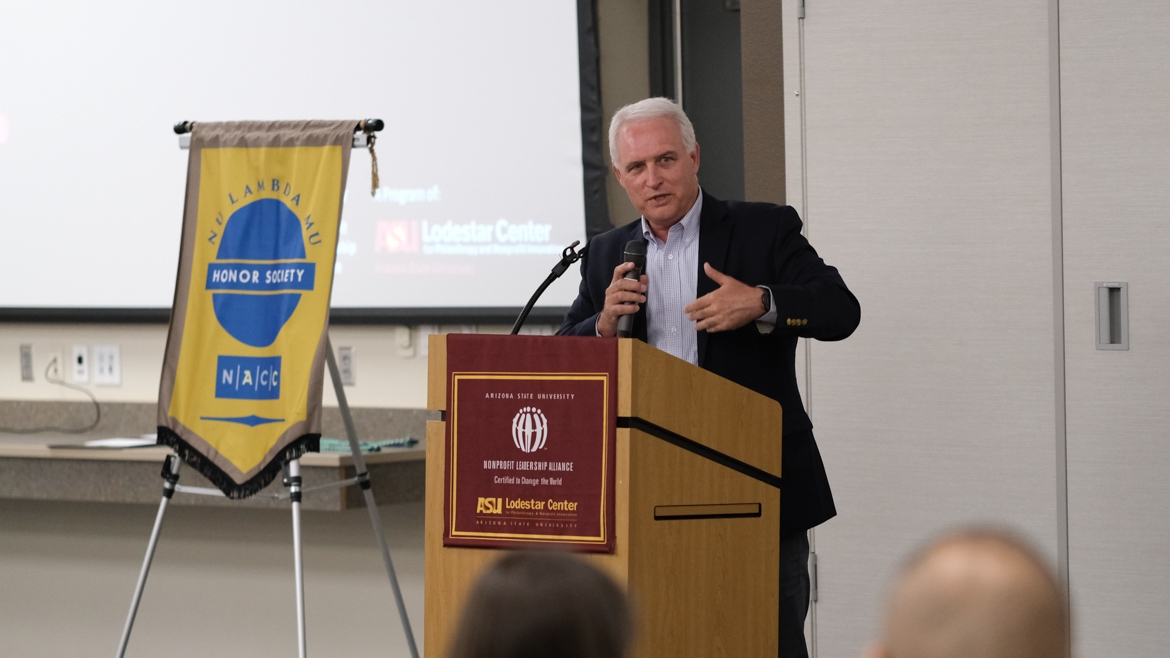 Standing behind a podium on stage, Robert Ashcraft, executive director of the ASU Lodestar Center for Philanthropy and Nonprofit Innovation, addresses the audience. Behind him is a projector screen and a small banner reading "Nu Lambda Mu, Honor Society, NACC." On the podium is a maroon and gold banner reading "Nonprofit Leadership Alliance", alongside the logo for ASU and ASU Lodestar Center.