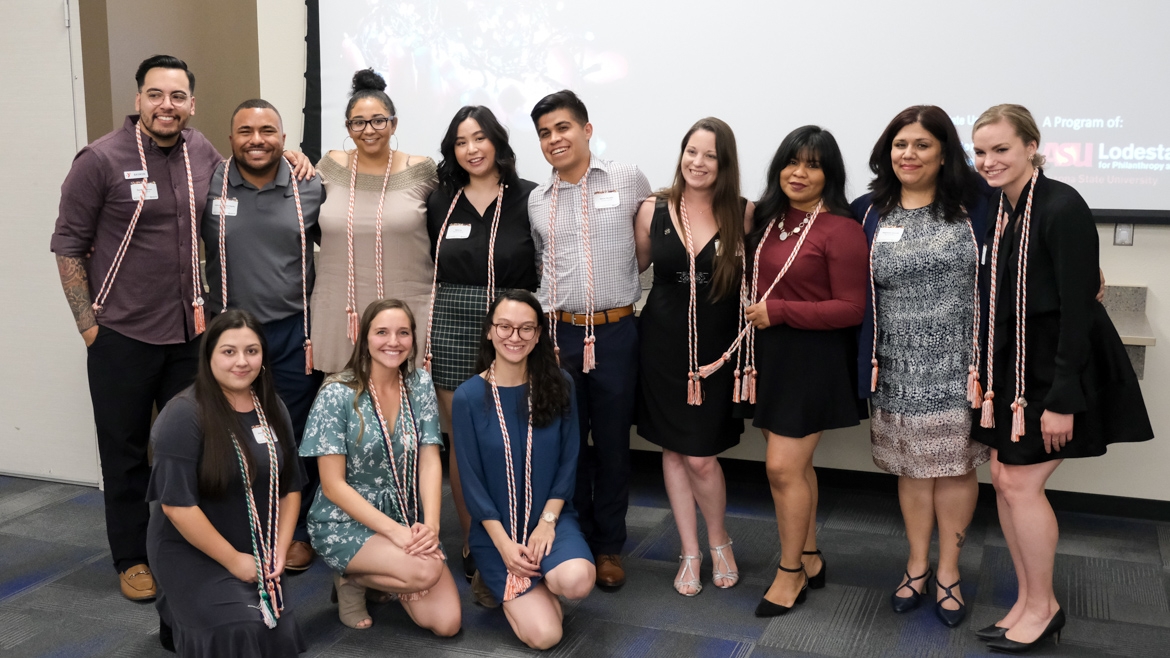 Standing in front of a projector screen, Nonprofit Leadership Alliance Student Association graduating seniors of 2019 pose together and smile. Each person has a tassle drapped over their shoulders.