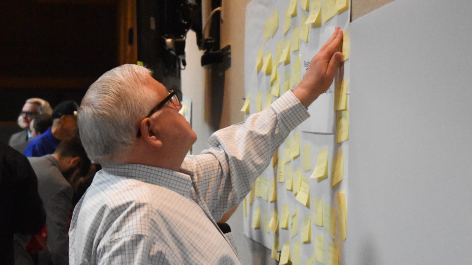 An elderly person stands and adds a yellow post-it note to several others on a wall.