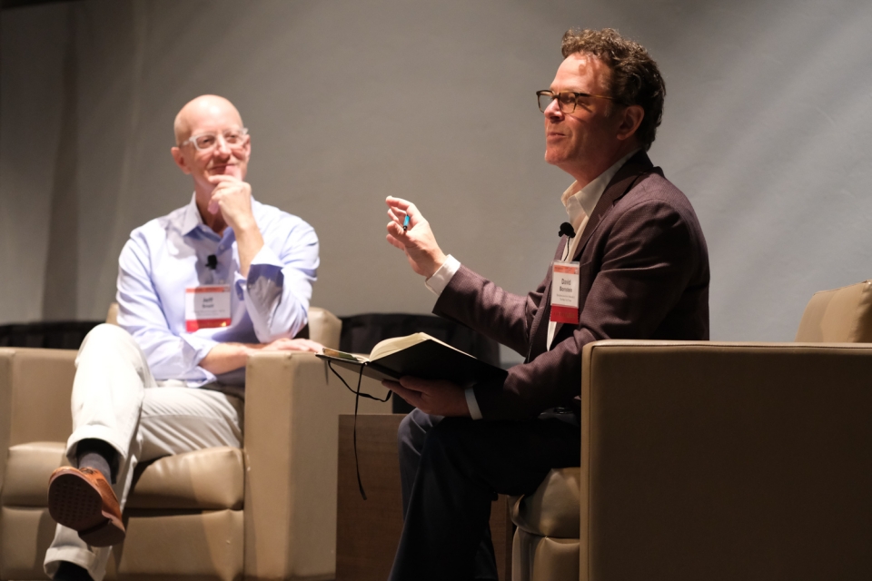 On a stage, 2 people (Jeff Snell on the left, and David Bornstein on the right) sit in comfy armchairs and discuss the Solutions Collaboratory.