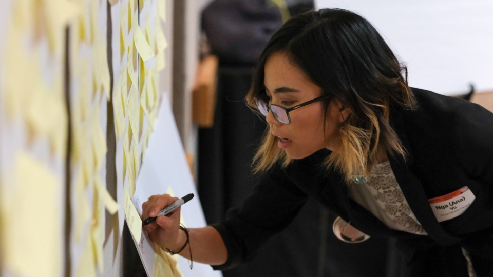 A young person wearing glasses leans in to write on a board leaning against a wall full of yellow post-it notes.