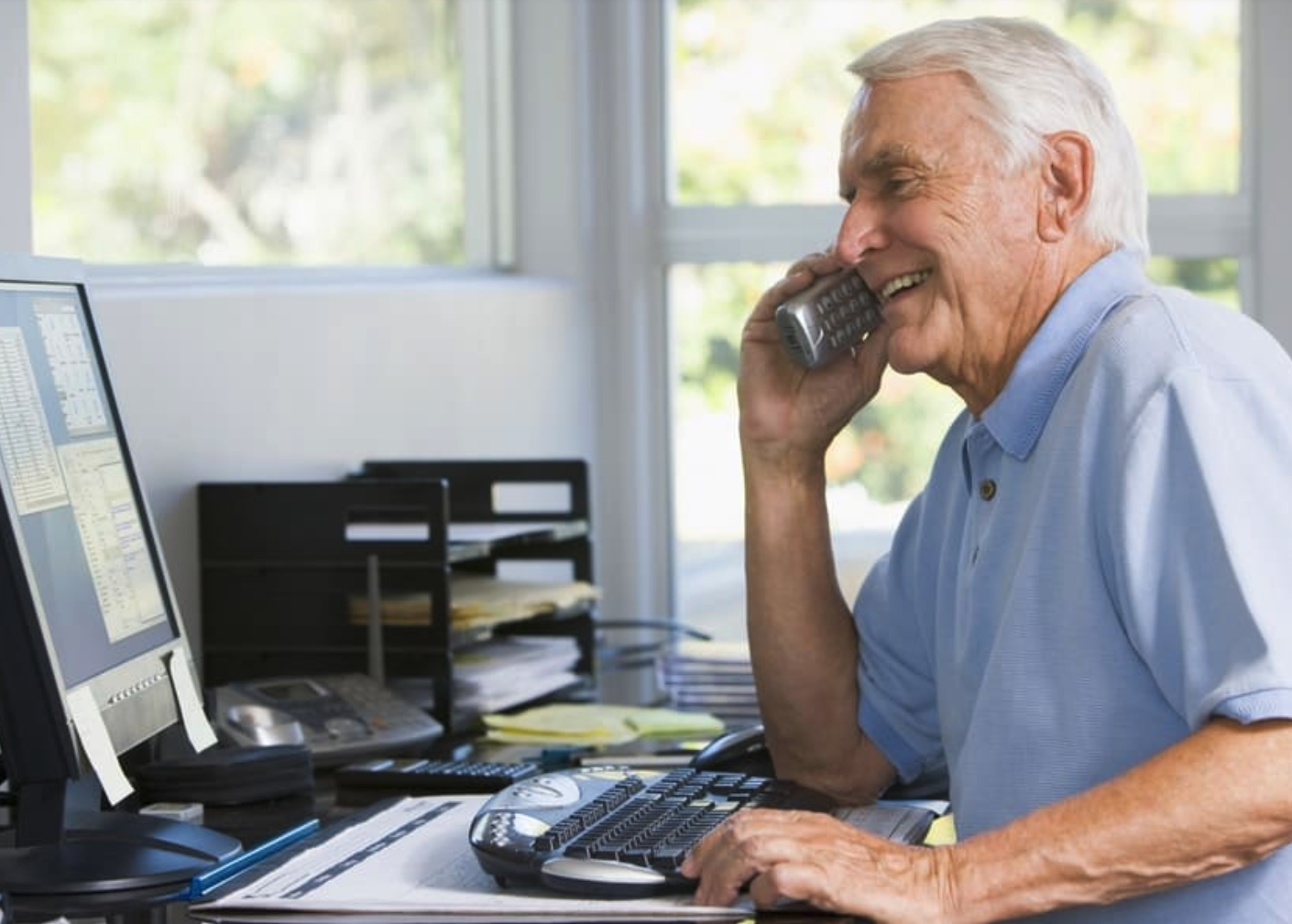 A senior citizen sits at a desk and smiles while on a phone call.