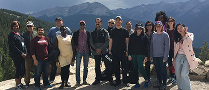 Participants in the Fellowship program gather at the top fo a mountain. In the background are distant trees and purple mountains against an overcast sky.