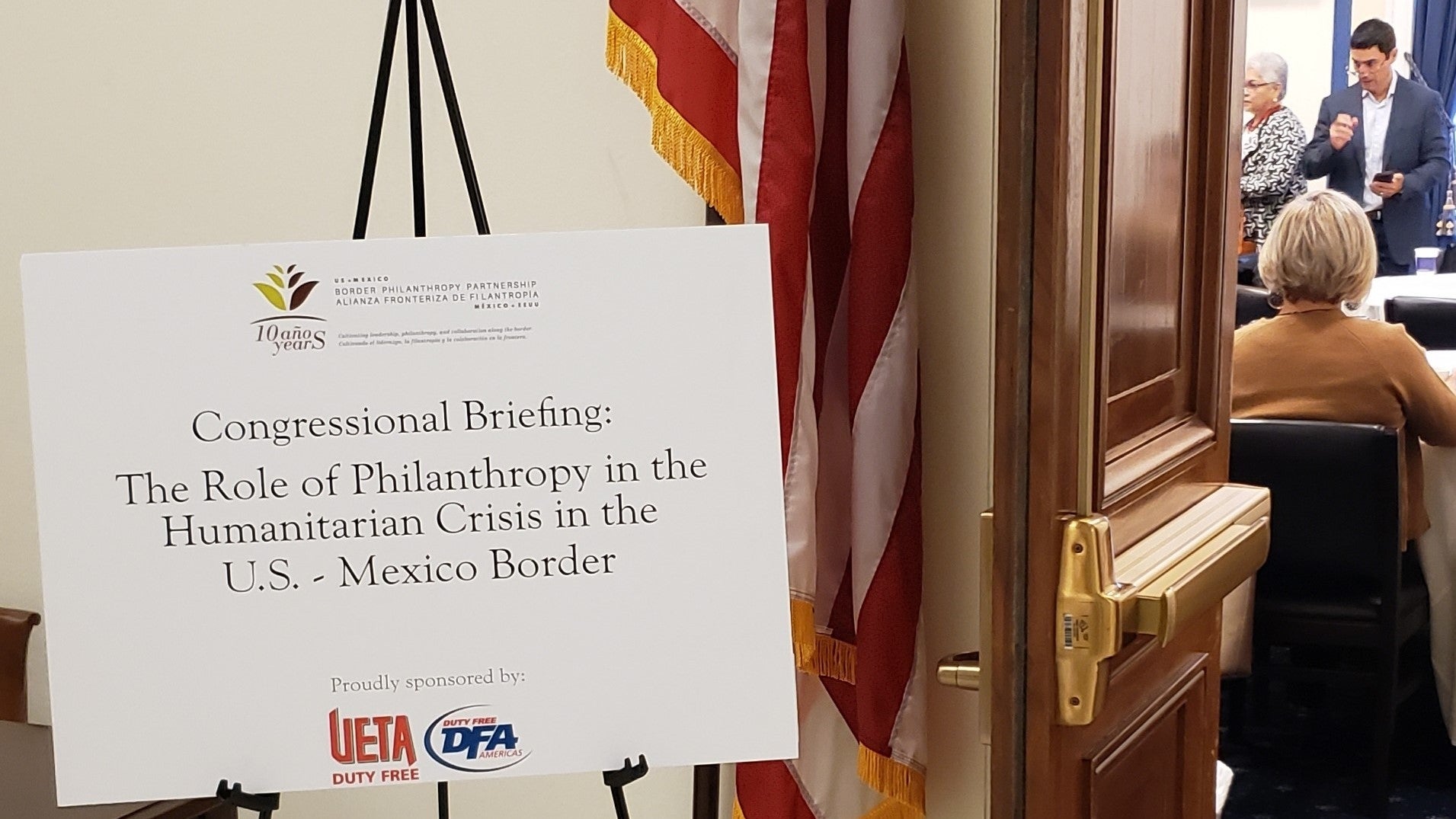 A sign outside of an open conference room reads "Congressional Briefing: The Role of Philanthrophy in the Humanitarian Crisis in the U.S.-Mexican Border". There are several logos included on the sign.