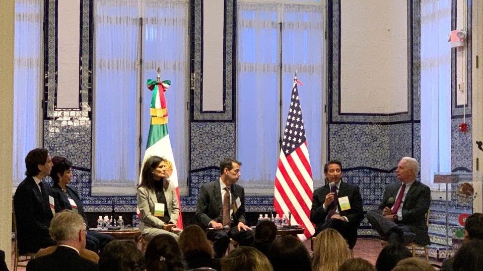 6 people sit on a stage in front of a large audience. On the stage are an American and Mexican flag.