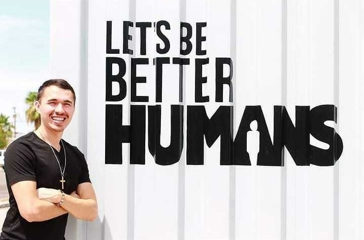 Carlitos Hernandez crosses his arms and leans against a large wall with the phrase "Let's be better humans" written on the wall.