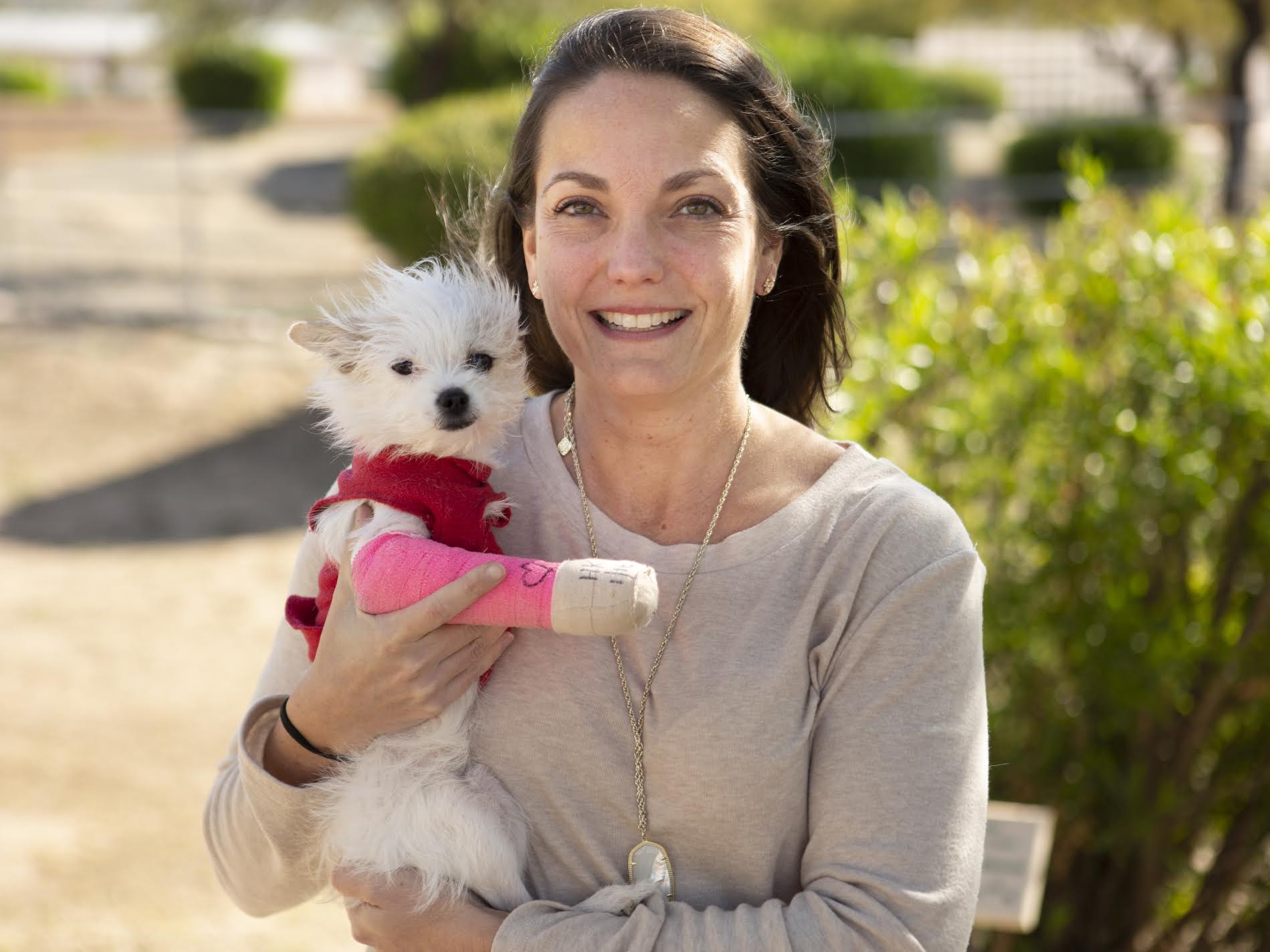 Carrie Hughes is outdoors and smiling while holding a small white dog that has a pink cast on its front right leg.
