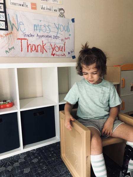 A kid with a leg brace on their left calf sits in a chair. Behind them are cubbies and a handwritten sign reading "We miss you!". The sign is hand-signed by several people.