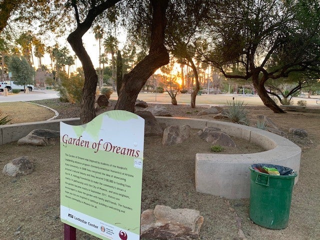 A park with a "Garden of dreams" sign. The park is mainly gravel ground, and in the background there are palm trees, a sunset, and a green grass field.