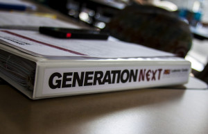 A binder lies on a table. The binder spine says "Generation Next"