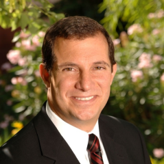 A professional photo portrait of John Scola wearing a suit. In the background are plants.
