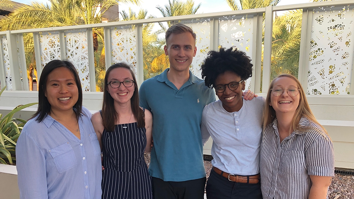 Members of the Nonprofit Leadership Alliance Student Association stand side-by-side while outdoors. Behind them is a decorate white fence and palm trees.
