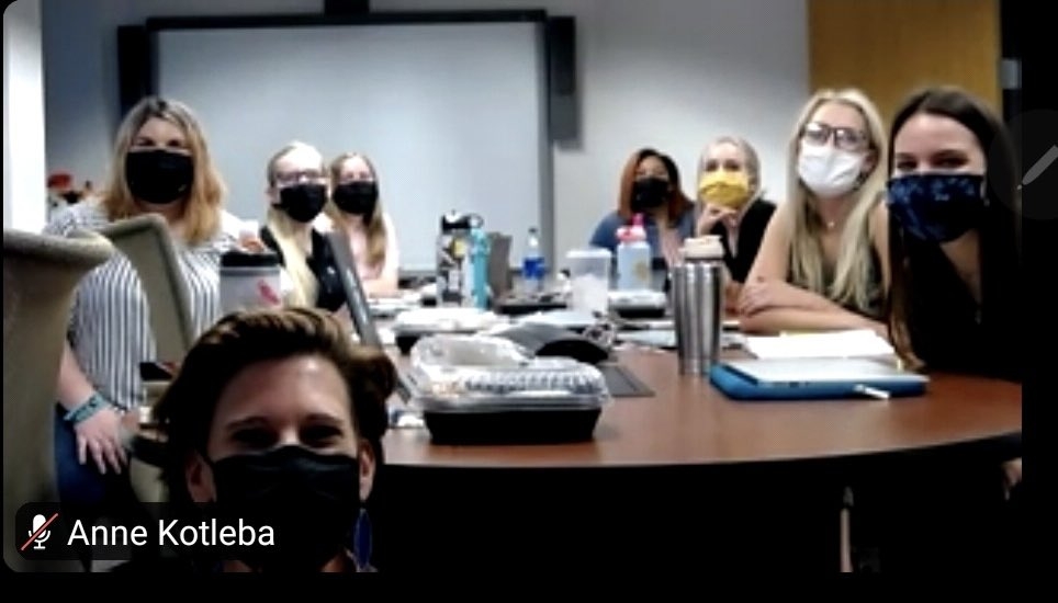 People wearing face masks sit at a conference table. There are packaged snacks on the table.
