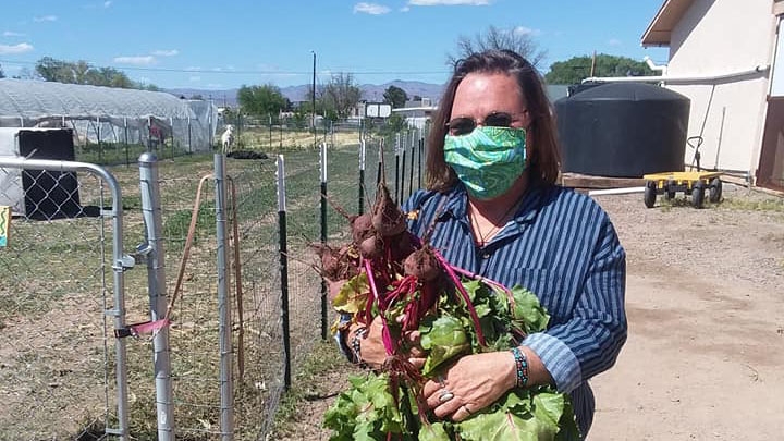A person wearing a face mask carries fresh plants. Behind them is a house, fence, and greenhouse.