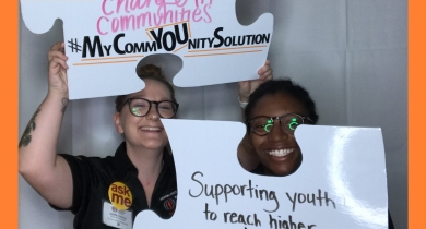 2 people smile and hold up white paper puzzle pieces. On the puzzle pieces are written phrases: "Supporting youth to reach higher" and "Changing Communities,  #MyCommYOUnitySolution".