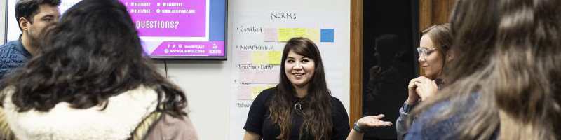 Reyna Montoya speaks to a couple other people while standing in a classroom with a projection screen and whiteboard with notes behind her.