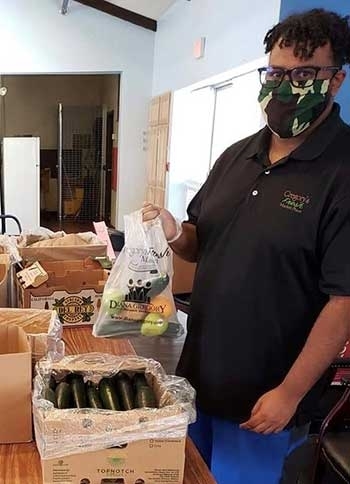 A person wearing a face mask holds up a plastic bag of vegetables. In front of them is a table with several boxes of food.