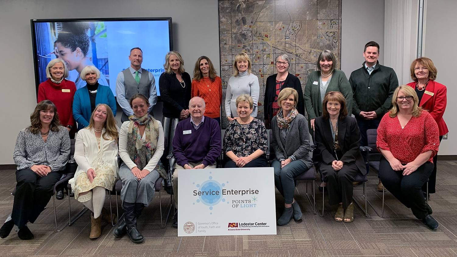 Members of the Arizona Service Enterprise Initiative's 17th cohort stand and sit in chairs inside a conference room. Behind them is a large monitor and map. In front of them is a sign reading "Service Enterprise, Points of Light" with multiple logos.