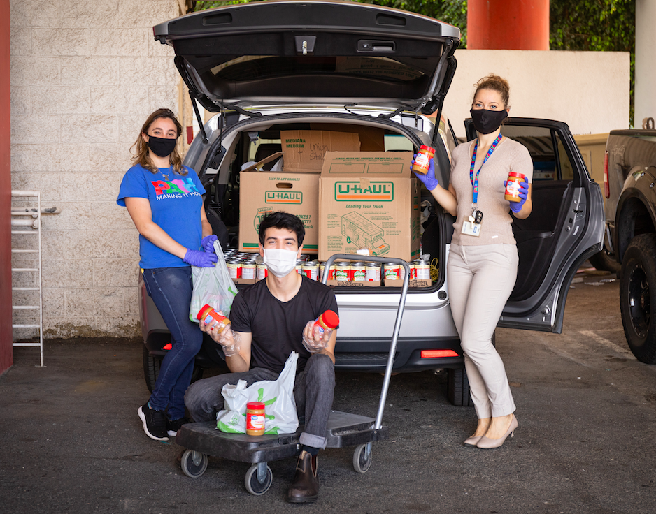In a parking lot, 3 people wearing face maks pause while putting canned food and boxes in a car trunk.
