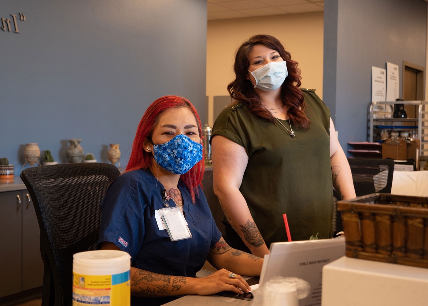 2 people wearing face masks are behind a desk within an office space.