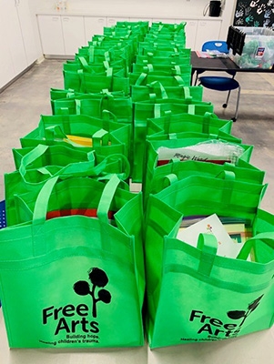 Green bags marked "Free Arts" are filled with items and lined up a table.