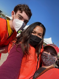 3 people, all wearing face masks and orange vests, smile.