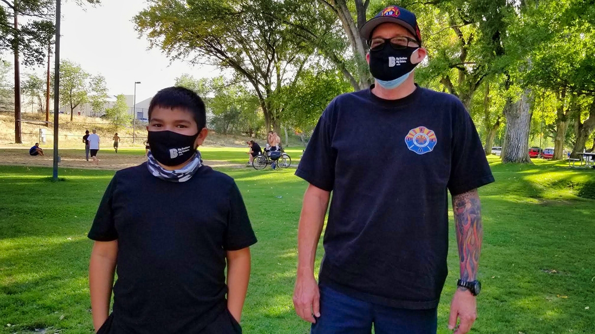 A kid and young adult both wear masks in a park. Behind them are trees, grass, and a gravel trail.