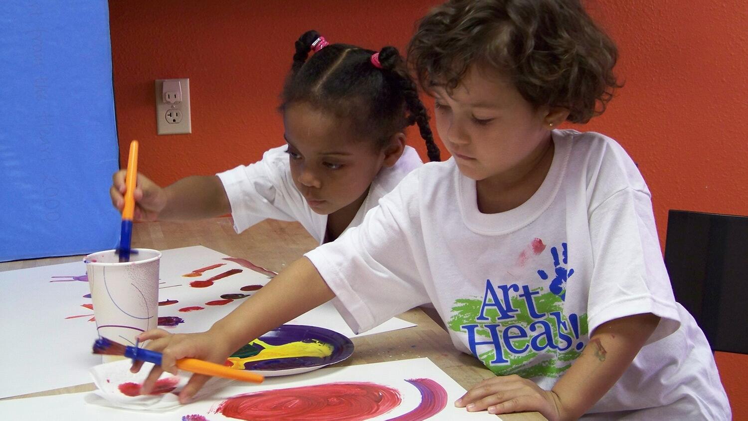 2 kids sit at a table and paint pictures.